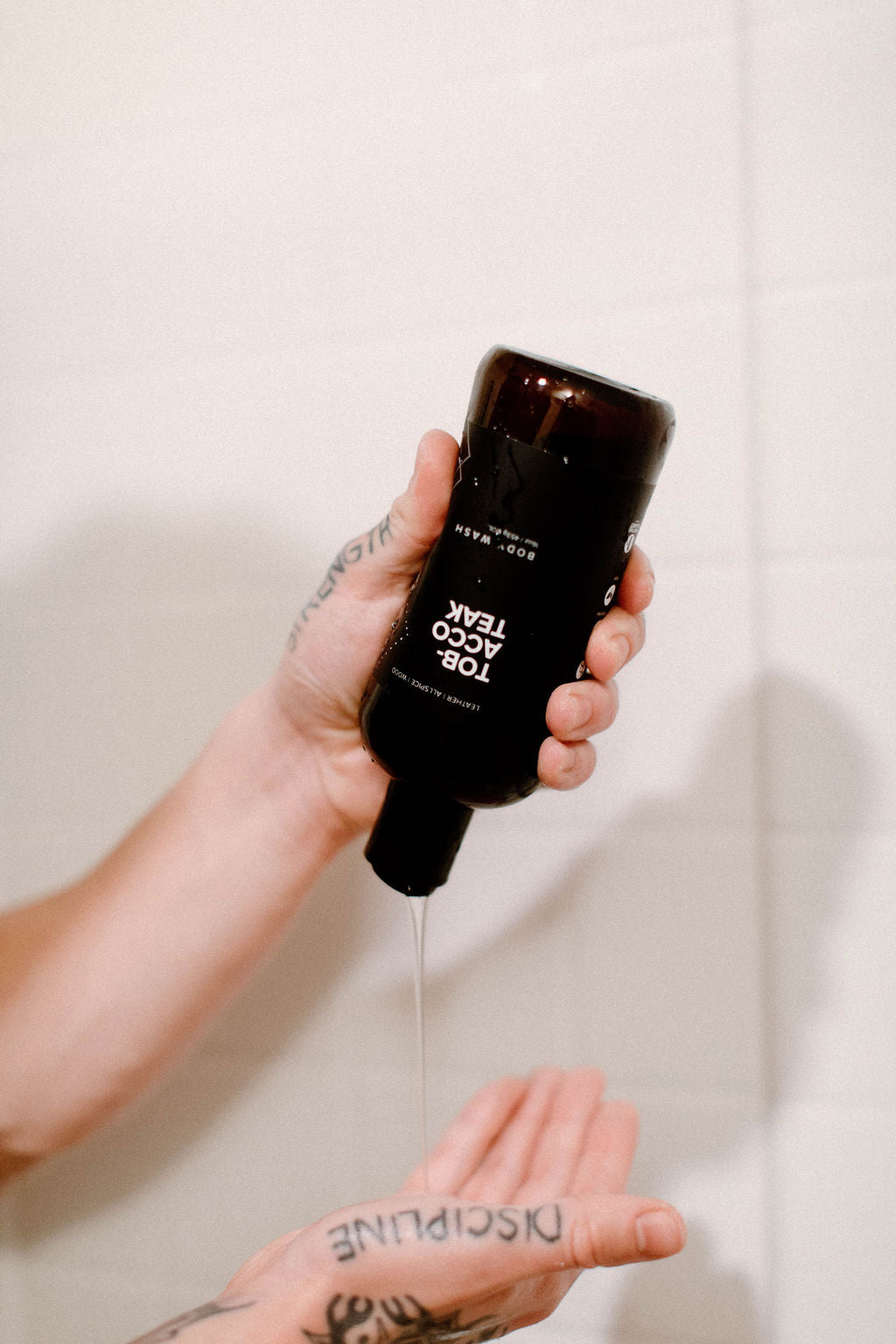 16oz Tobacco Teak Body Wash being dispensed into a hand in a white tiled shower. 