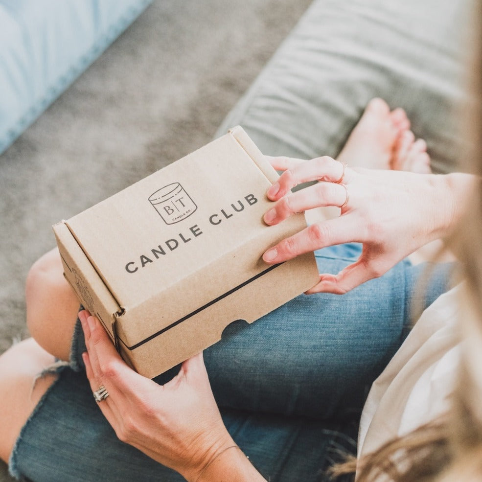 Candle Club shipping box being opened by female sitting on couch. 