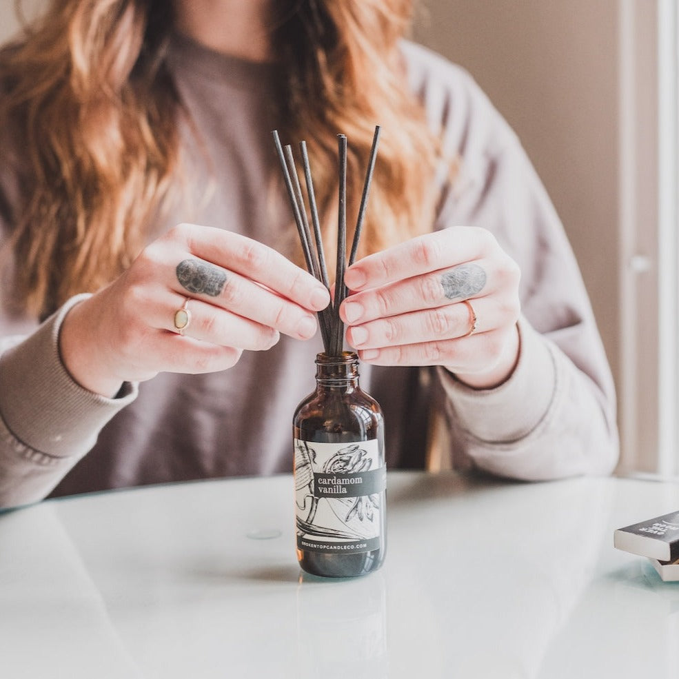 Cardamom Vanilla Reed Diffuser on a table with woman's hands adjusting the reeds. 