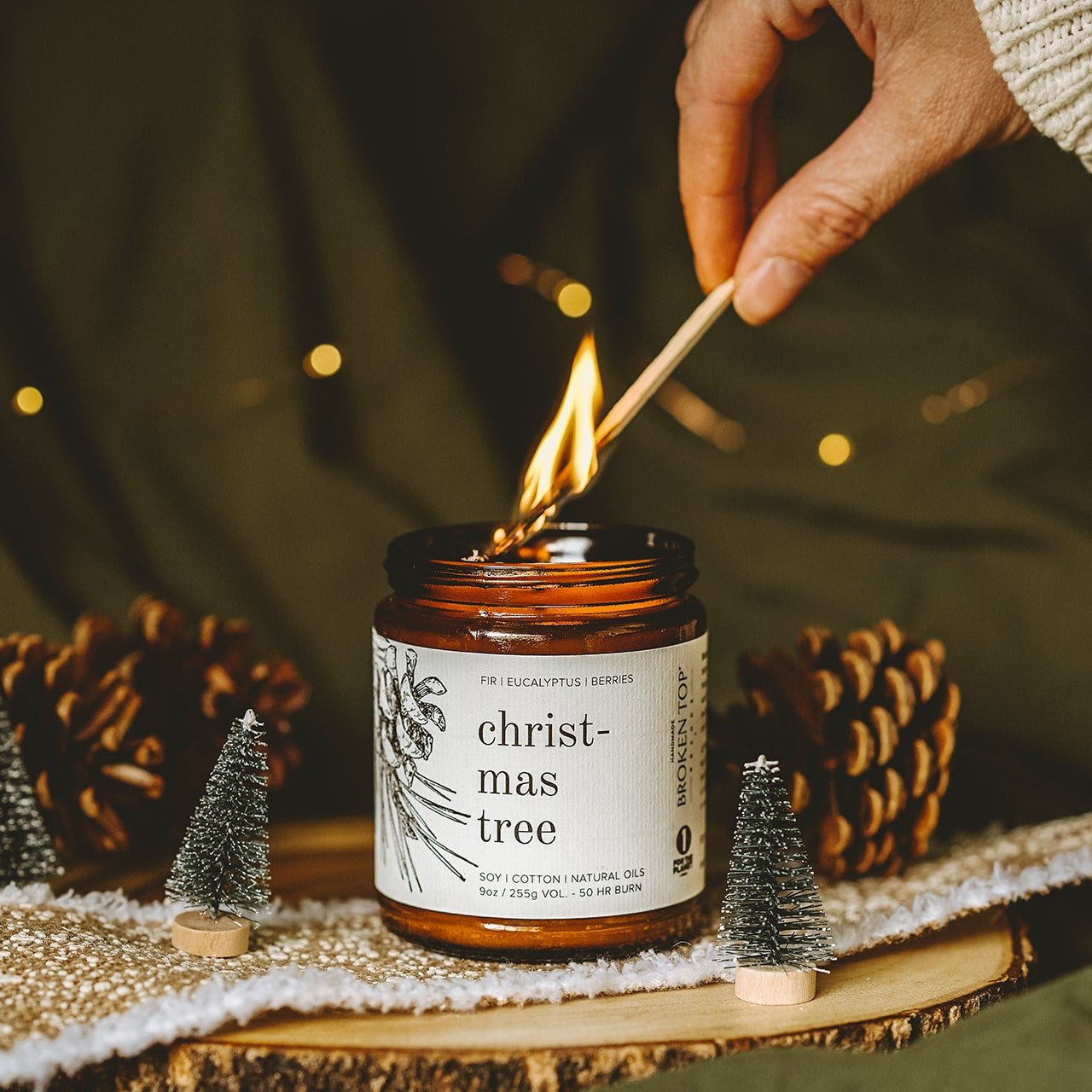 9oz Christmas Tree Soy Candle being lit by a match next to small Christmas decor. 