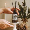 Christmas Tree Reed Diffuser being positioned by two hands on a white stand next to two pinecones. 