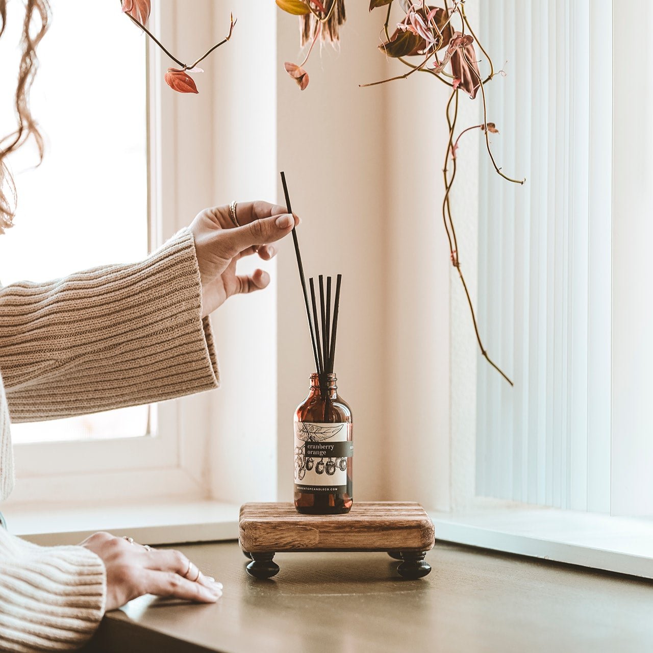 Cranberry Orange Reed Diffuser on a small wooden shelf being adjusted by woman's hands. 