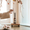 Cranberry Orange Reed Diffuser on a small wooden shelf being adjusted by woman's hands. 