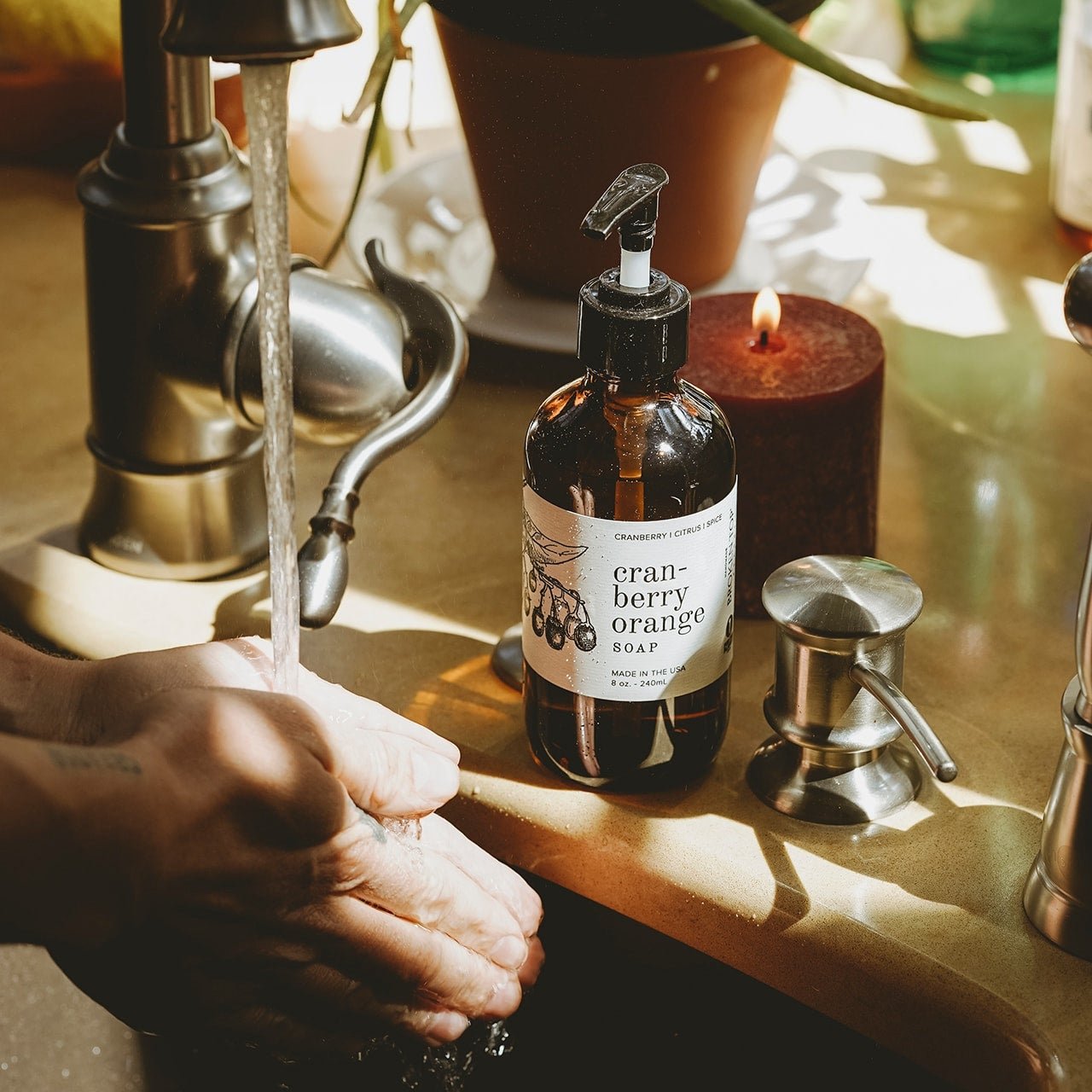 8oz Cranberry Orange Liquid Soap next to a sink with hands washing under running faucet. 