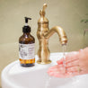 Mount Bachelor 8oz Liquid Soap on the sill of a bathroom sink with hands washing under running water. 