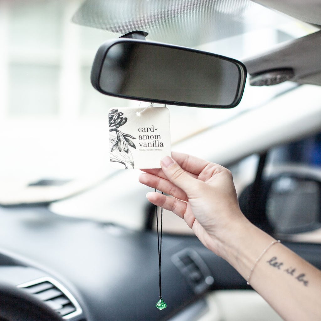 Cardamom Vanilla Car Freshener hanging from rearview mirror with hand grasping it. 