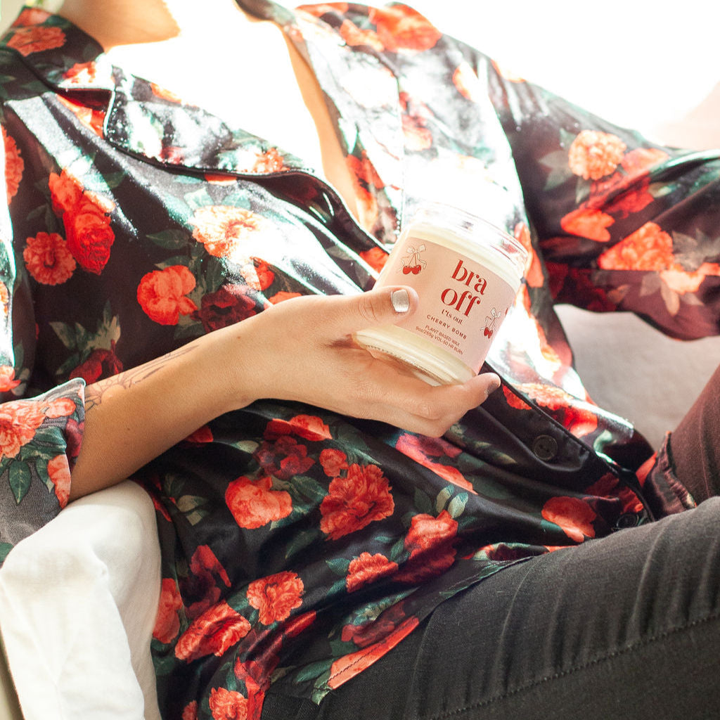 9oz 'Bra Off T*ts Out' Soy Candle being held by a woman wearing a floral silk shirt. 