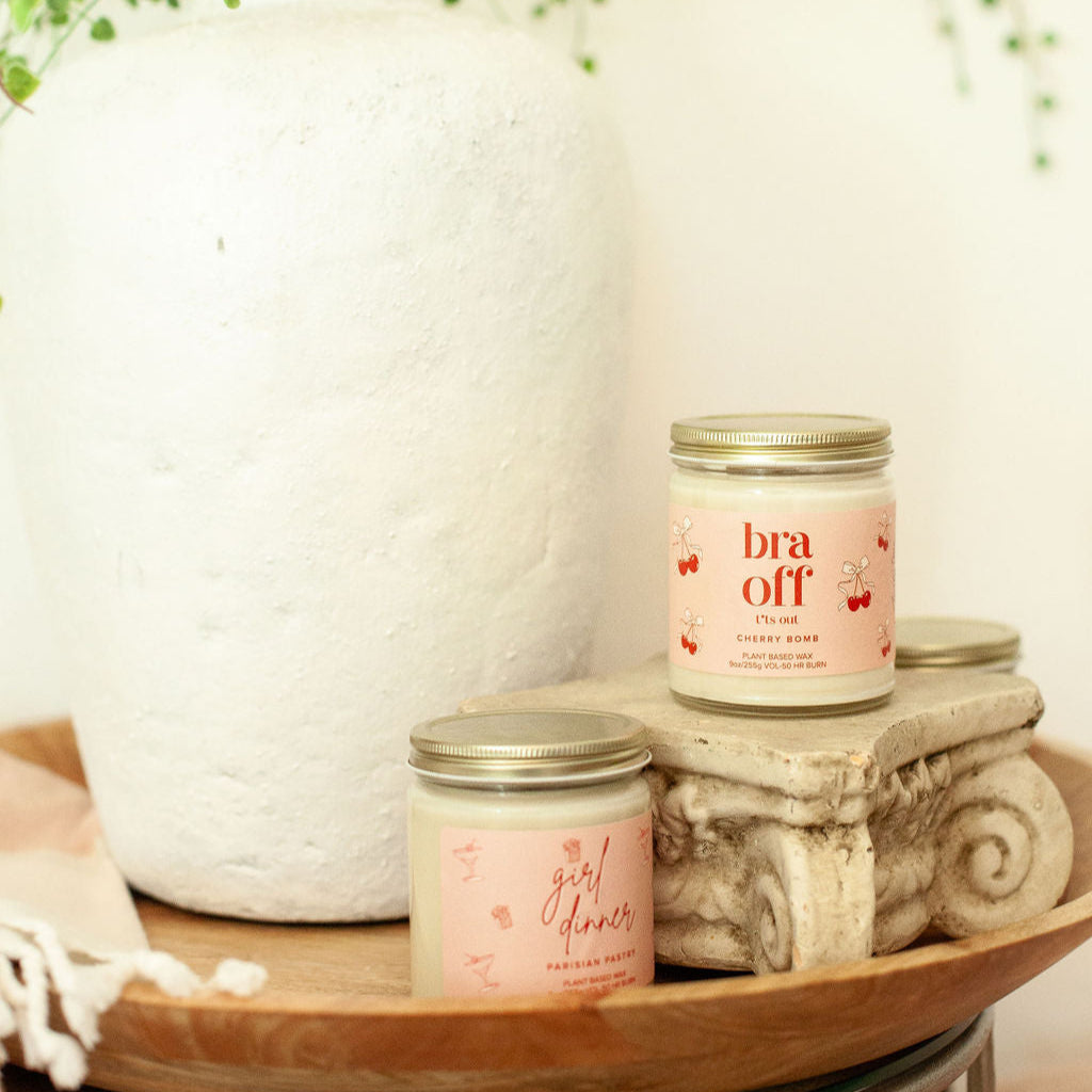 9oz 'Bra Off T*ts Out' Soy Candle on a small pedestal on a side table in front of a large house plant. 