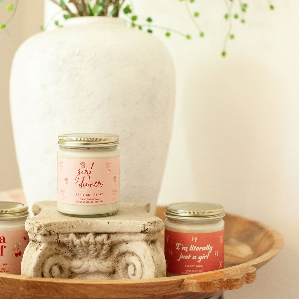 9oz 'Girl Dinner' Soy Candle on small pedestal on wooden side table in front of a large house plant. 