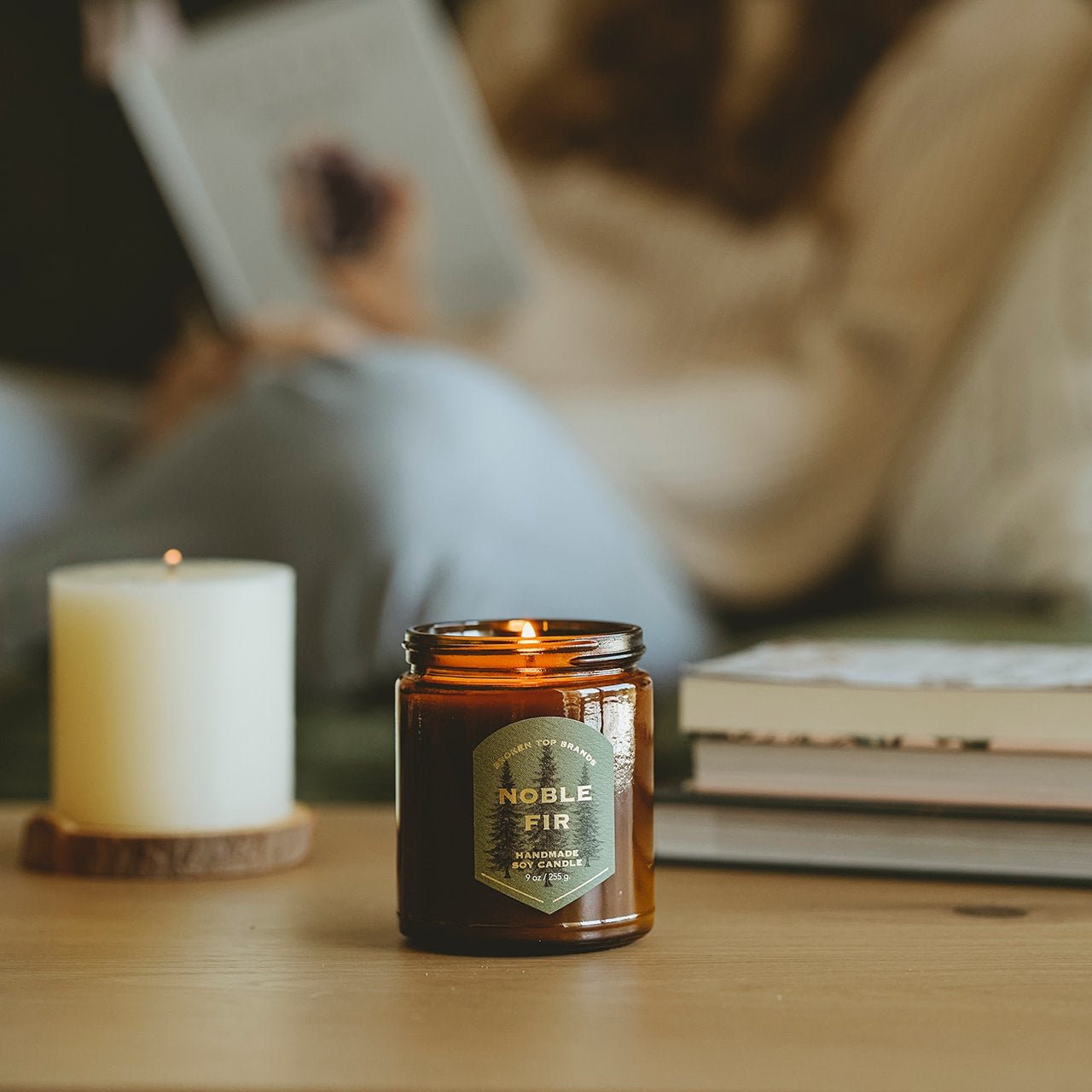 9oz Noble Fir Soy Candle on a coffee table with a woman reading in the background. 