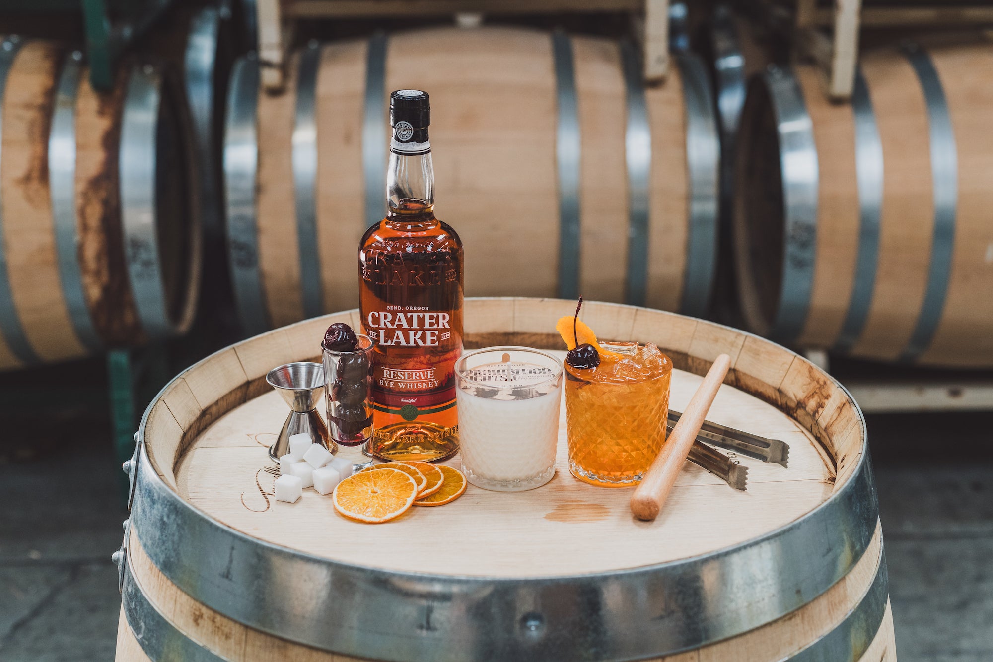 11oz Old Fashioned Soy Candle on a barrel next to Crater Lake Whiskey, sliced oranges, cherries, sugar cubes, and an old fashioned cocktail. 