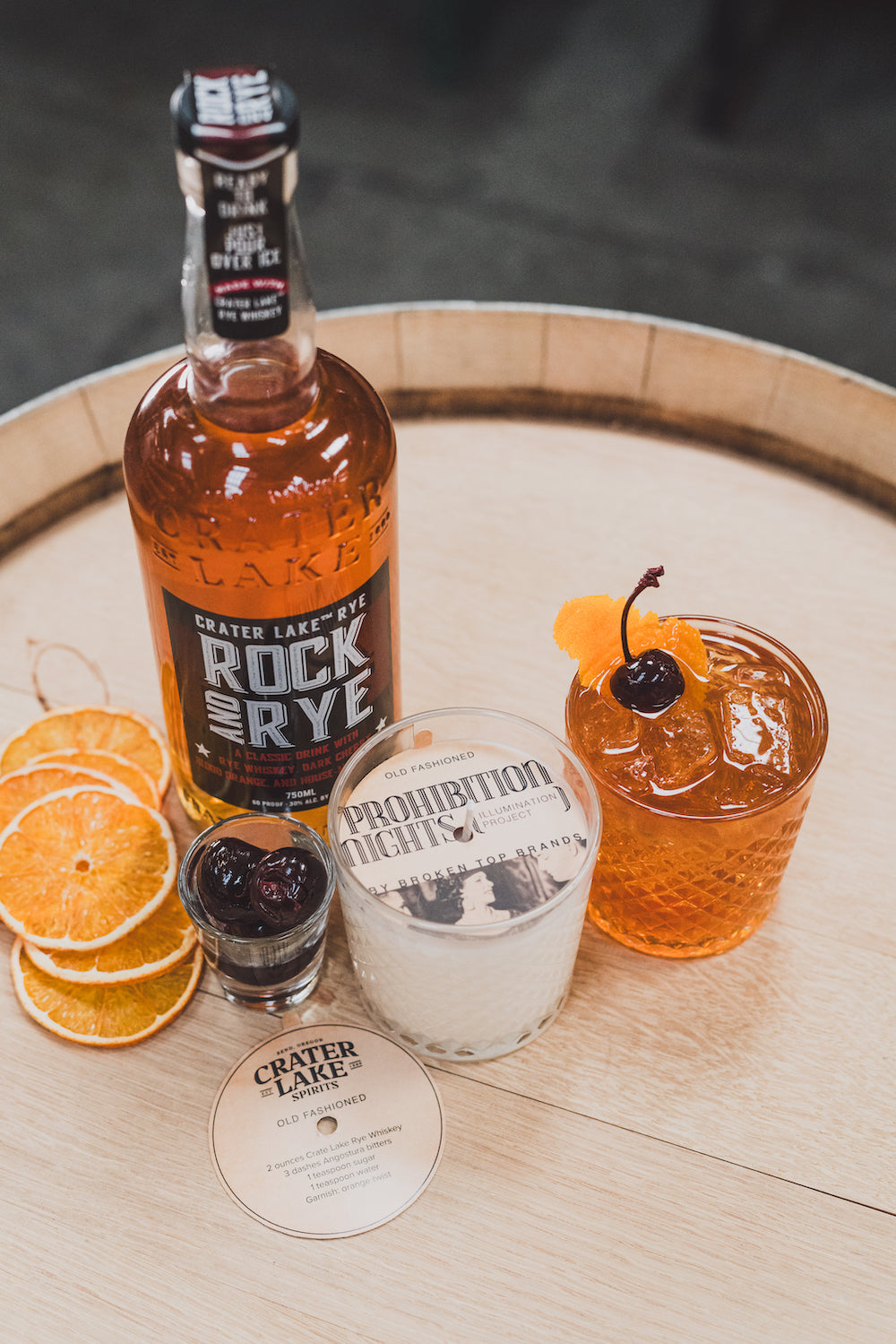 11oz Old Fashioned Soy Candle on a barrel with Crater Lake Whiskey, orange slices, cherries, and an old fashioned cocktail. 